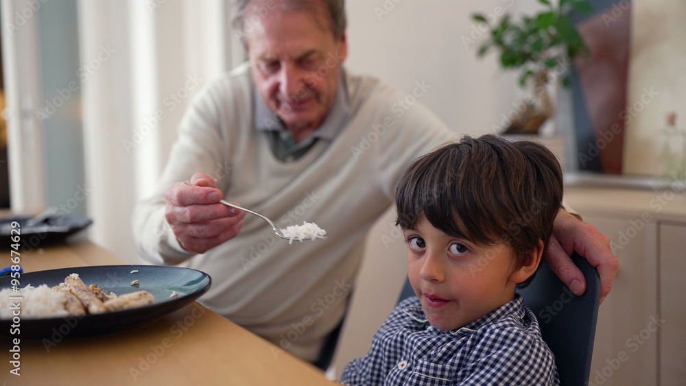 Fototapeta premium Elderly man feeding a young boy at a dining table, showcasing intergenerational care and bonding during mealtime in a cozy home environment