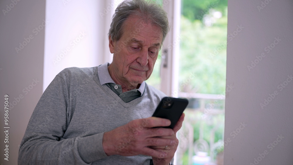 Elderly man sitting by a window, focused on his smartphone. Sunlight streams in, highlighting his contemplative expression in a serene, green background Elderly man sitting by a window, focused on his smartphone. Sunlight streams in, highlighting his contemplative expression in a serene, green background