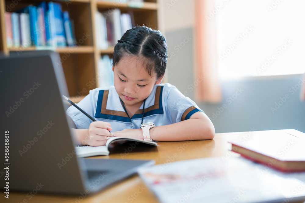 Young girl in a school uniform studying alone at a desk, writing in a notebook while referencing a laptop, smiling and focused, surrounded by bookshelves, capturing a moment of independent learning Young girl in a school uniform studying alone at a desk, writing in a notebook while referencing a laptop, smiling and focused, surrounded by bookshelves, capturing a moment of independent learning