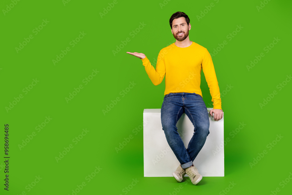 Full size photo of pretty young male hold empty space sit cube wear trendy yellow outfit isolated on green color background Full size photo of pretty young male hold empty space sit cube wear trendy yellow outfit isolated on green color background