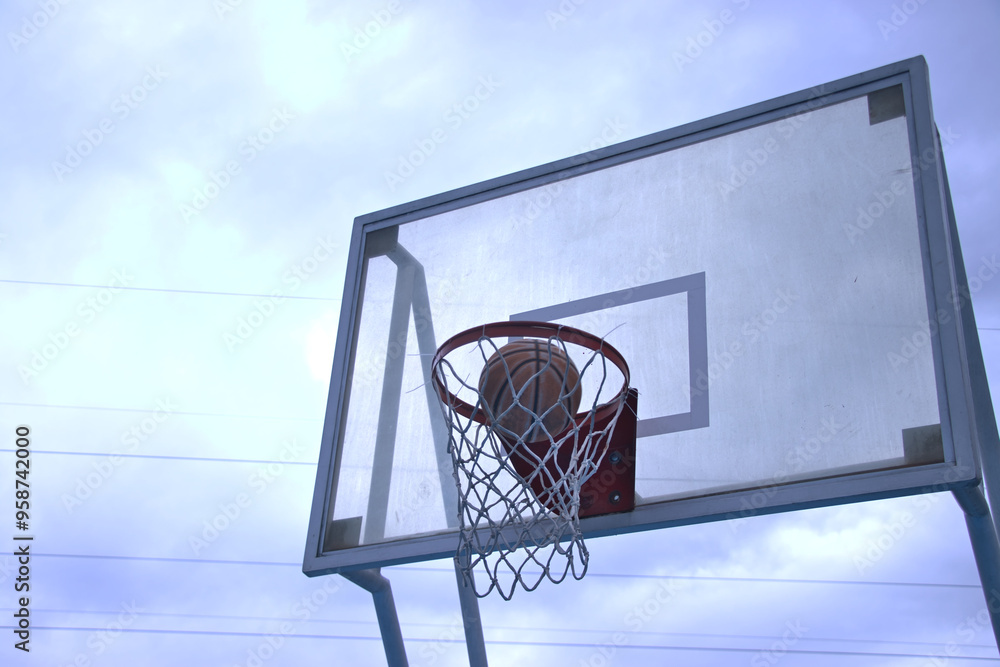 A basketball is halfway through the hoop after a successful shot during an outdoor game on a cloudy day. The image captures the moment of scoring, emphasizing the accuracy and timing of the play A basketball is halfway through the hoop after a successful shot during an outdoor game on a cloudy day. The image captures the moment of scoring, emphasizing the accuracy and timing of the play