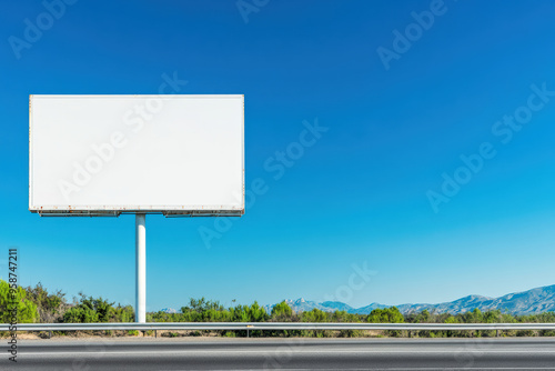 A blank billboard stands next to a highway with a clear blue sky and distant mountains, ideal for outdoor advertising in a scenic area.