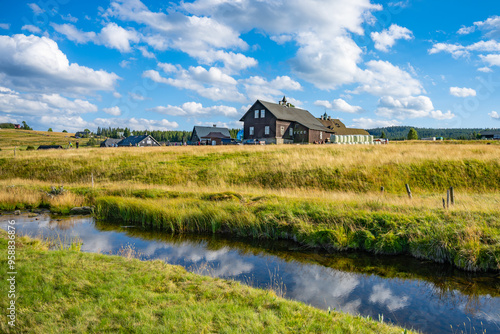 A picturesque view of Jizerka village in the Jizera Mountains showcases quaint wooden houses under a bright blue sky. Lush green fields and a reflective stream enhance the tranquil summer landscape.