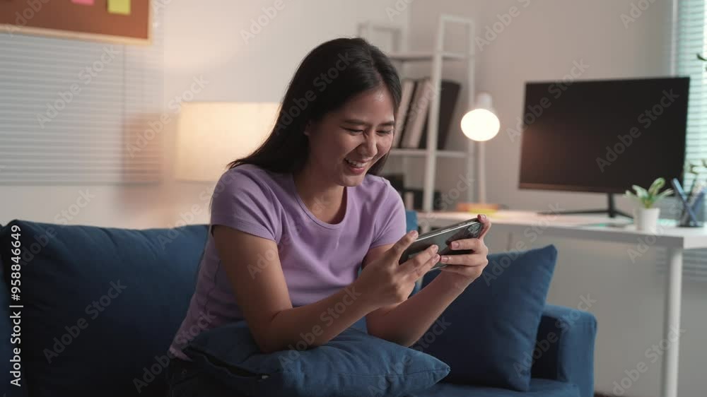 Young woman is playing a game on her phone while sitting on a sofa in her living room. She is happy and excited as she is winning Young woman is playing a game on her phone while sitting on a sofa in her living room. She is happy and excited as she is winning