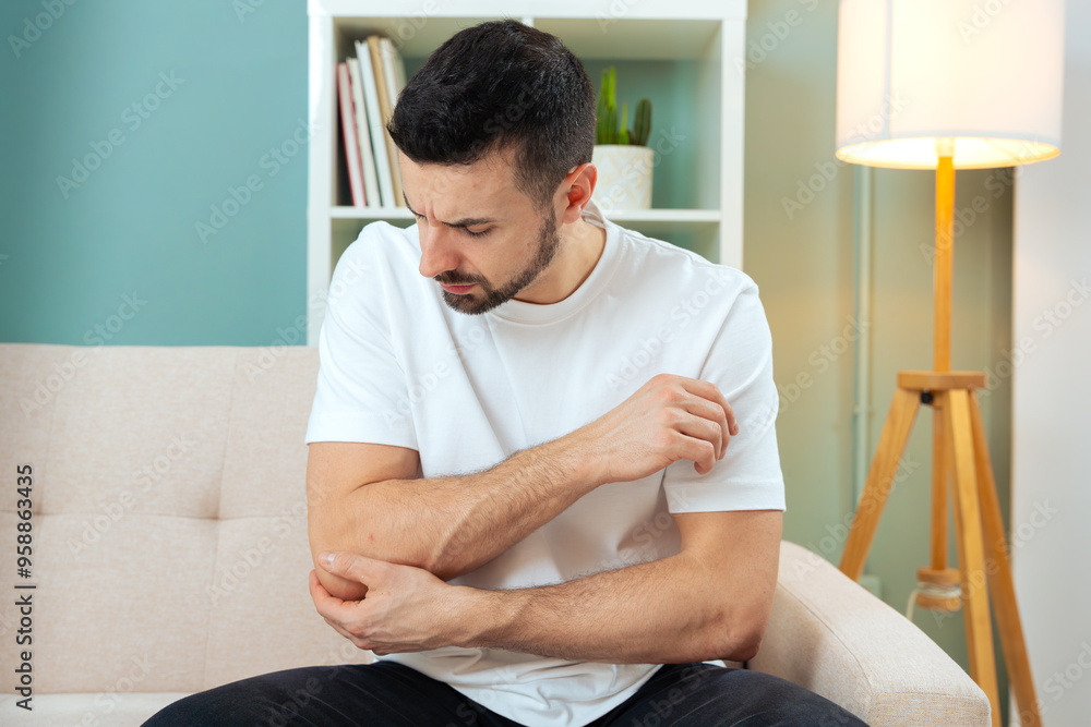 The man is sitting on a gray sofa and holding his arm. Concept of medicine and health, muscle problems. The man suffers from elbow pain, bone problems. Arm pain, suffering and pain. The man is sitting on a gray sofa and holding his arm. Concept of medicine and health, muscle problems. The man suffers from elbow pain, bone problems. Arm pain, suffering and pain.