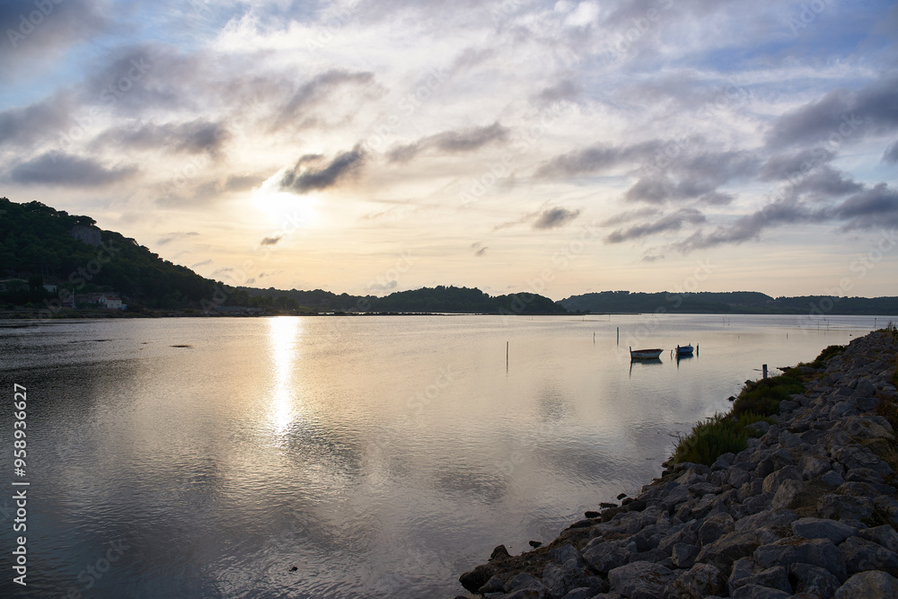 Sunset on a calm lake with the sun rising between the mountains with the sky covered by clouds and a small wooden boat in the background with its reflection in the water Sunset on a calm lake with the sun rising between the mountains with the sky covered by clouds and a small wooden boat in the background with its reflection in the water