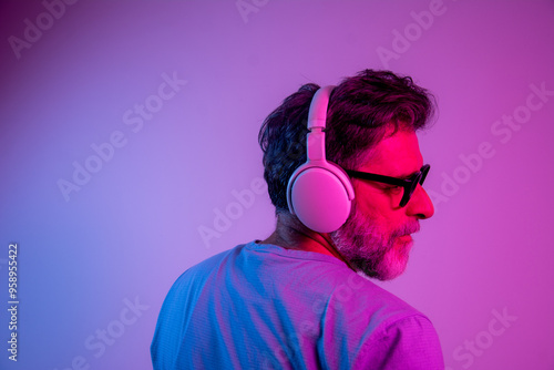 Enjoying his favorite music. Happy senior stylish man glasses with headphones listening and smiling while standing against blue neon background photo