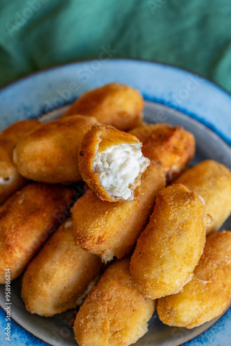 A vibrant blue plate filled with delicious, golden-brown croquettes resting on a soft green fabric background. One croquette is split open, revealing the creamy and rich bechamel filling inside, showc