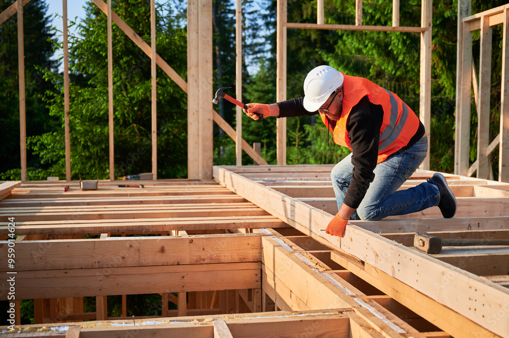 Carpenter erecting a two-story, timber-framed dwelling close to the woods. A bespectacled and bearded man is hammering nails into the structure, donning a safety helmet and a construction vest. Carpenter erecting a two-story, timber-framed dwelling close to the woods. A bespectacled and bearded man is hammering nails into the structure, donning a safety helmet and a construction vest.