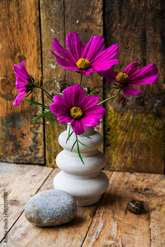 A vase with purple flowers sits on a wooden table beside a rock. The flowers are arranged in a way that they are all facing the same direction, creating a sense of harmony and balance