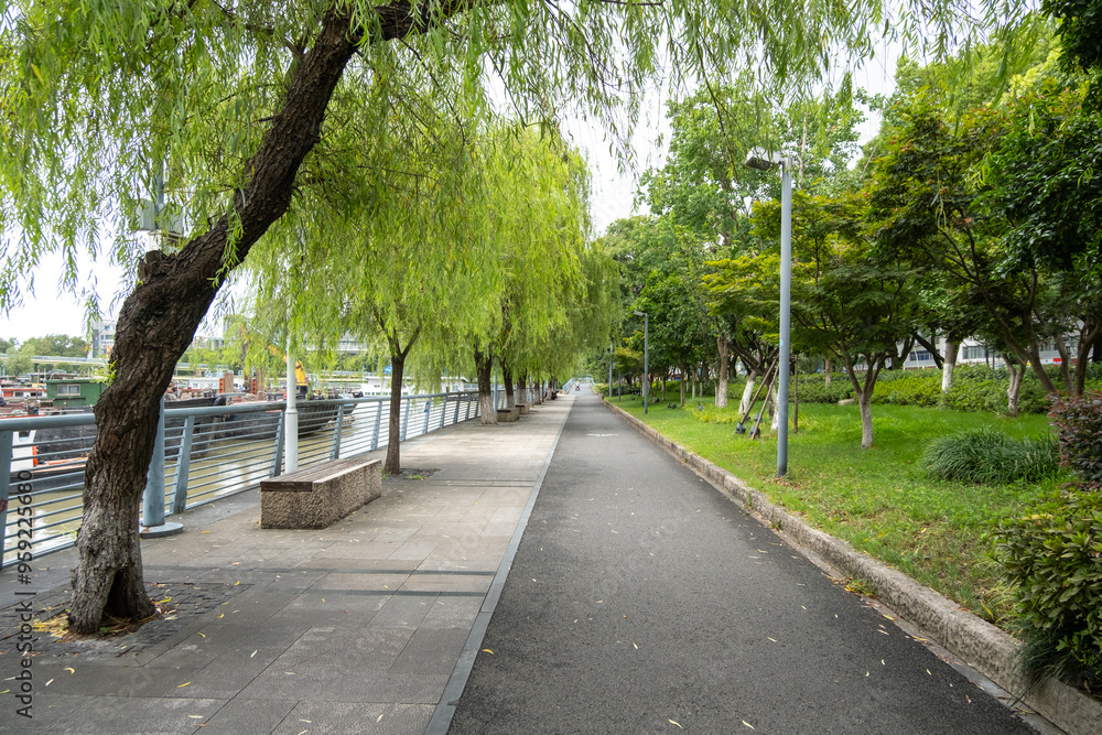 Obraz premium Background texture of a scenic walkway along a riverbank, lined with lush green trees,including weeping willows, providing shade. An urban park with a pedestrian footpath and cycling lane.Ningbo China