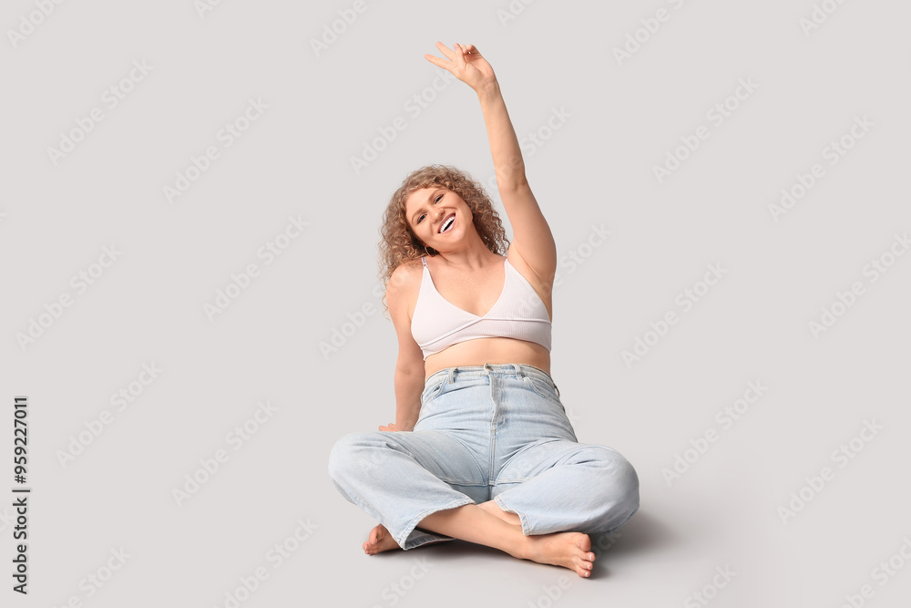Beautiful young happy body positive woman in stylish jeans sitting on grey background Beautiful young happy body positive woman in stylish jeans sitting on grey background