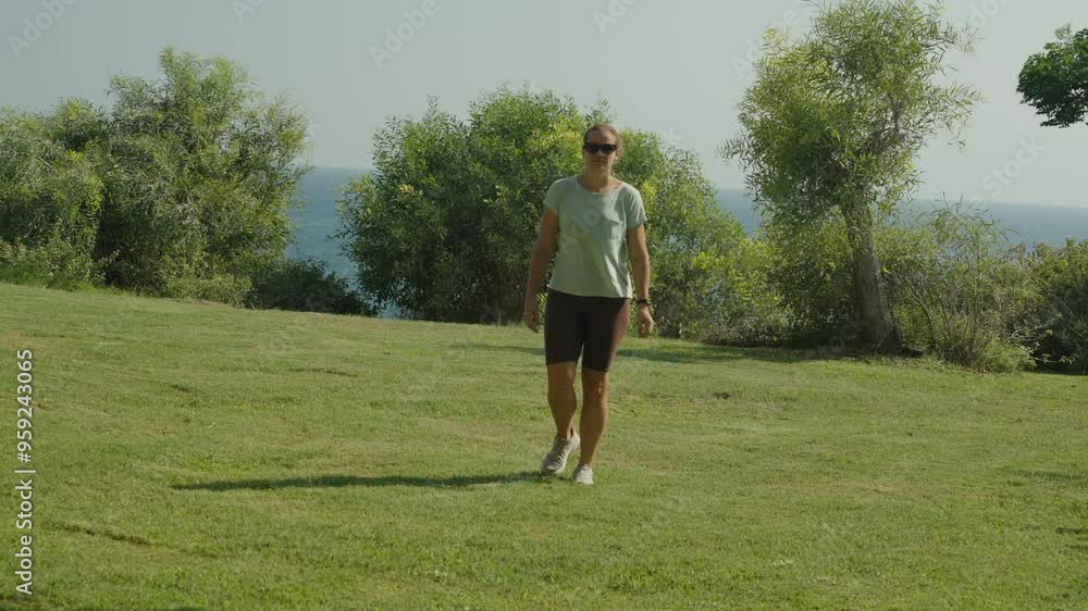 Zoom out, Front View, Woman Walking Across Grassy Field, Surrounded by Trees and Bushes, Ocean in the Background, Bright and Tranquil Day. Zoom out, Front View, Woman Walking Across Grassy Field, Surrounded by Trees and Bushes, Ocean in the Background, Bright and Tranquil Day.
