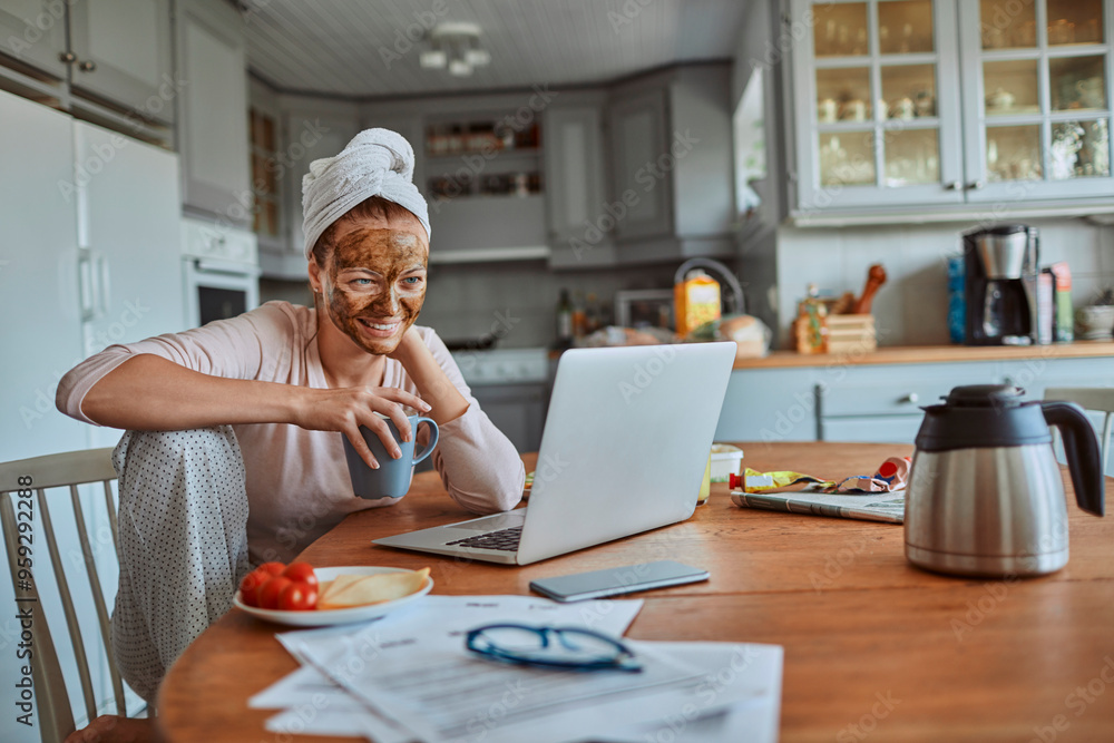 Woman with face mask enjoying breakfast and coffee while working on laptop in kitchen Woman with face mask enjoying breakfast and coffee while working on laptop in kitchen