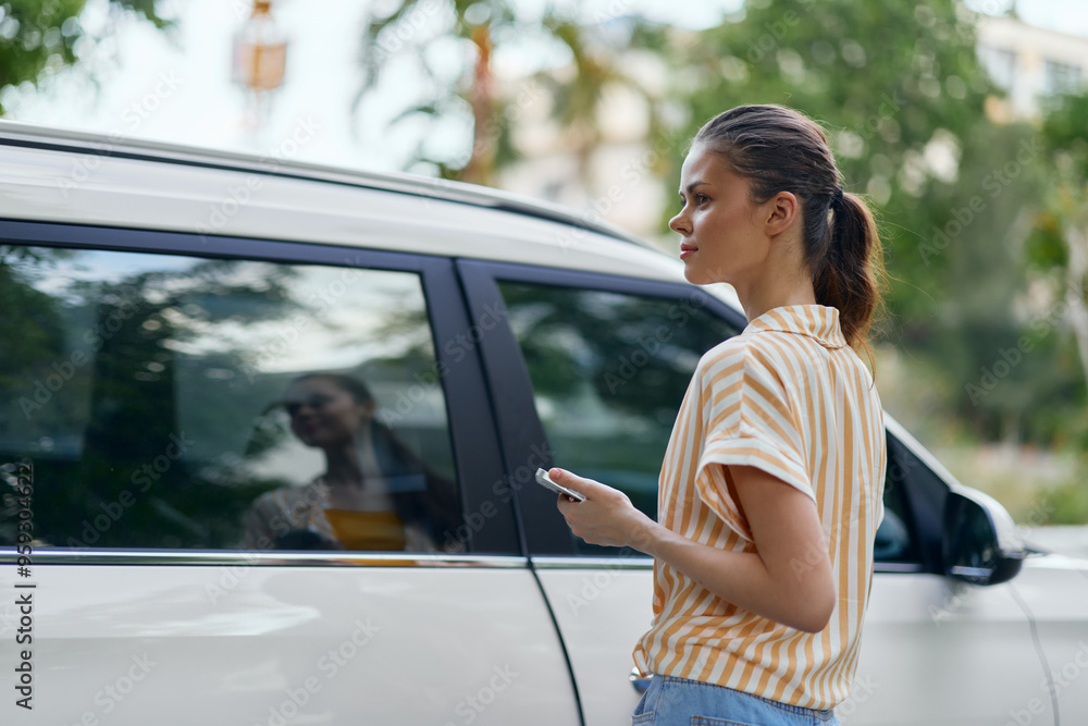 Woman standing near a white car, checking her phone, outdoors, surrounded by greenery and soft sunlight Casual attire and focused expression highlight modern lifestyle Woman standing near a white car, checking her phone, outdoors, surrounded by greenery and soft sunlight Casual attire and focused expression highlight modern lifestyle