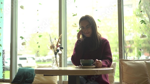 A young woman in glasses talking on the phone and drinking coffee in a cafe with a panoramic window.