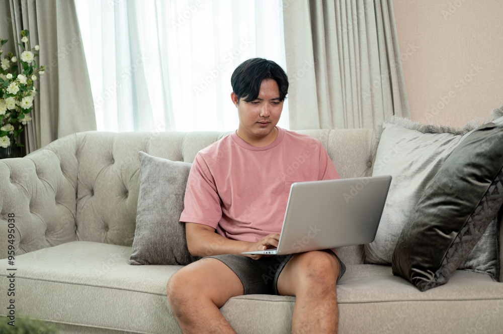 An Asian man in casual clothes using his laptop on a couch in the living room. An Asian man in casual clothes using his laptop on a couch in the living room.