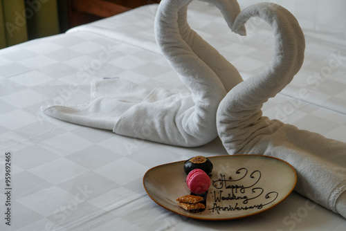Fototapeta Naklejka Na Ścianę i Meble -  Happy Wedding Anniversary. Two swans made from towel with a cake on a white bed in hotel room.