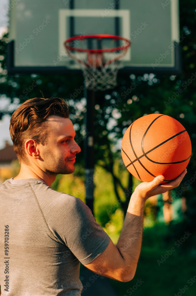 Man in a gray shirt holds a basketball in one hand while looking towards it, with a basketball hoop in the background. The outdoor scene is bright and vibrant, perfect for sports or leisure themes. Man in a gray shirt holds a basketball in one hand while looking towards it, with a basketball hoop in the background. The outdoor scene is bright and vibrant, perfect for sports or leisure themes.