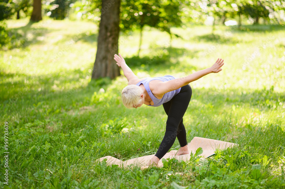Middle aged woman gracefully performs yoga pose standing barefoot on yoga mat laid out on soft grass in quiet park.Active lifestyle Middle aged woman gracefully performs yoga pose standing barefoot on yoga mat laid out on soft grass in quiet park.Active lifestyle