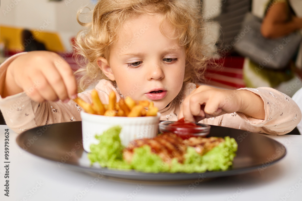 Unhealthy junk food for kids. Female toddler with wavy hair eating small snack at the table in cafe savoring the delicious fast meal with potatoes free and fried cutlet Unhealthy junk food for kids. Female toddler with wavy hair eating small snack at the table in cafe savoring the delicious fast meal with potatoes free and fried cutlet