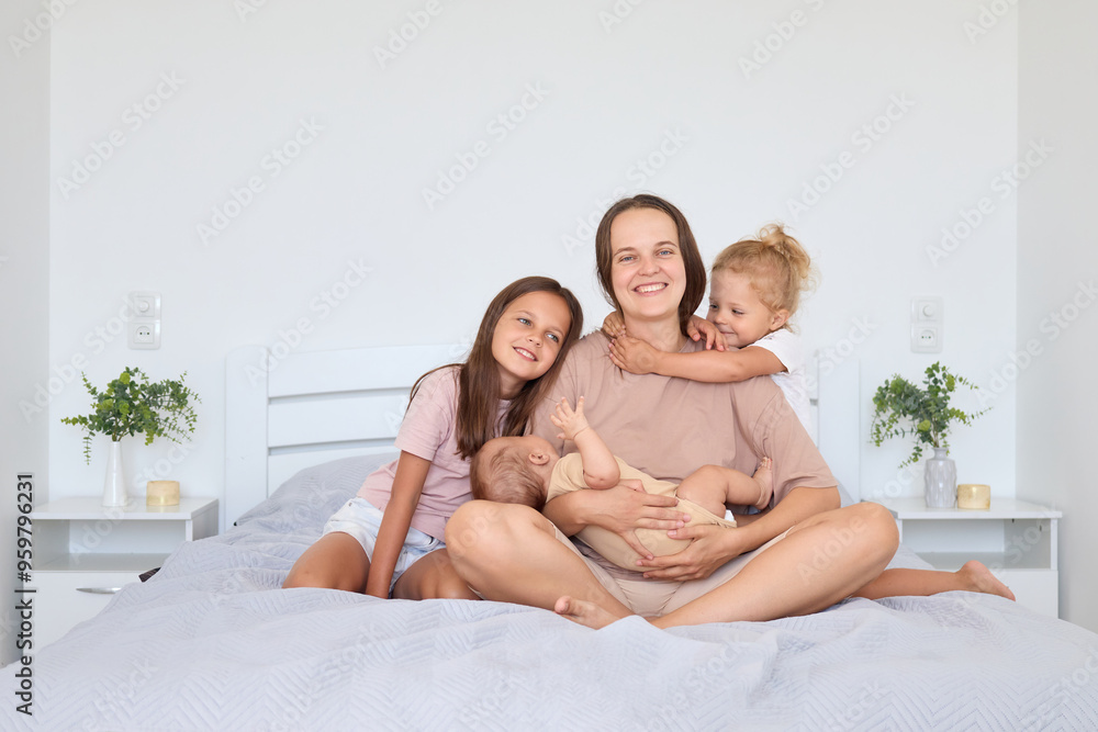 Happy mother sitting on bed with newborn baby and toddler daughters enjoying togetherness at home in light-filled bedroom joyful Caucasian woman with her charming little children Happy mother sitting on bed with newborn baby and toddler daughters enjoying togetherness at home in light-filled bedroom joyful Caucasian woman with her charming little children