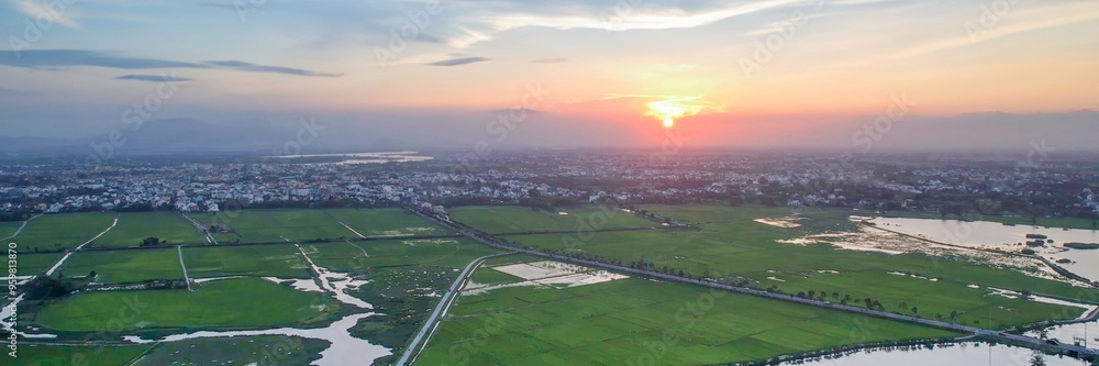 Fototapeta premium Aerial view of lush green rice fields and a peaceful town at sunrise, perfect for Asian rural landscapes and nature concepts