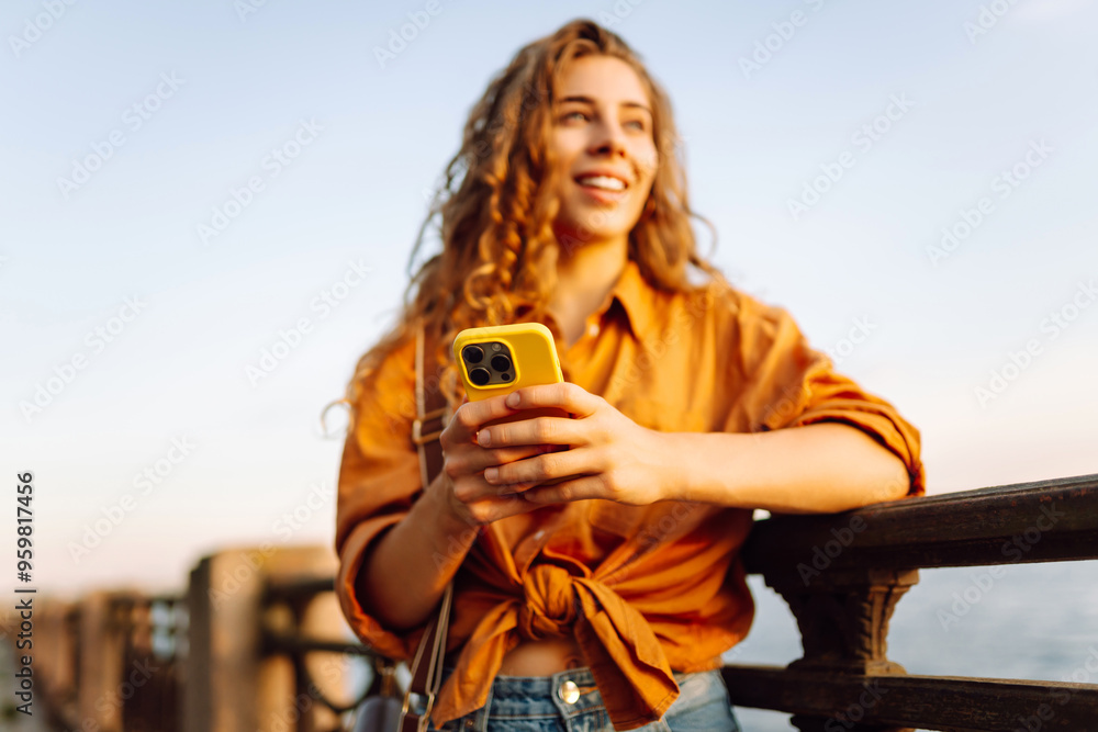 Fototapeta premium Young woman using smartphone by the waterfront during sunset, wearing a casual outfit while enjoying the warm evening light. Lifestyle, travel, nature, active life.