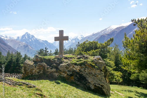 Stone cross with a bas relief of Christ on a huge moss-covered rock. Coniferous trees and snow capped mountains in the background