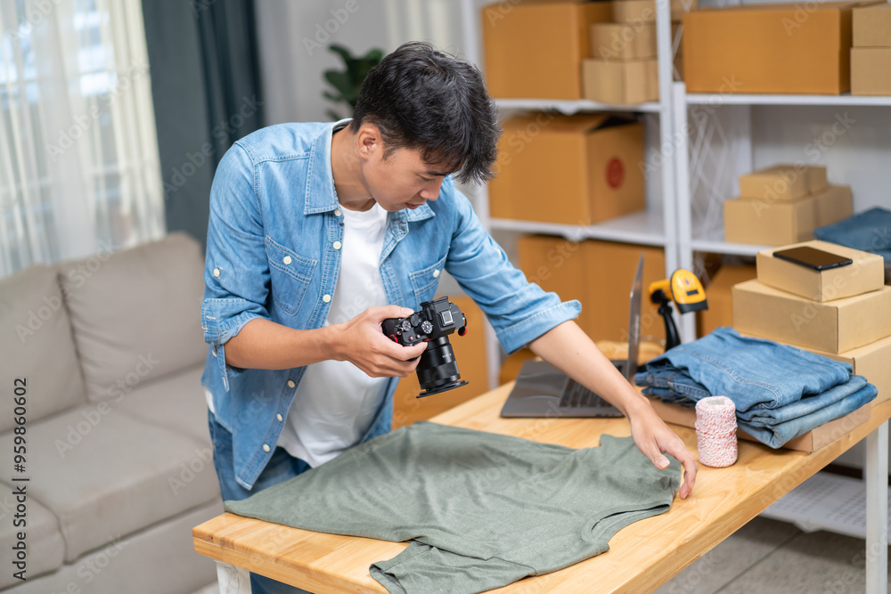 Young Asian man, distributor, online shop owner, small business owner, standing in warehouse, using camera to take pictures of products for sale.