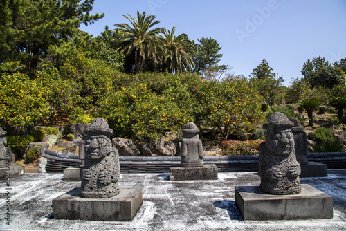 Seogwipo-si, Jeju-do, South Korea - March 23, 2021: Dolhareubangs at Yakcheonsa Temple against tangerine trees. Dolhareubang is grandfather made of stone and is believed to protect well-being photo