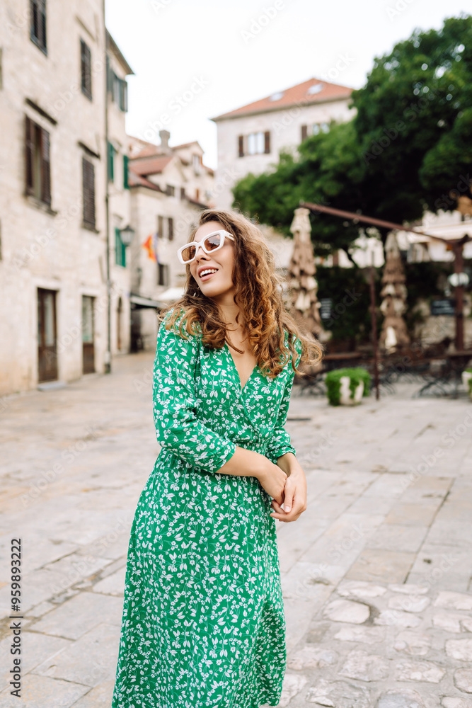 Naklejka premium A woman in a green dress enjoys a sunny day in the charming historic square of a Mediterranean town, gazing at the tall buildings and clock tower. Fashion, style, tourism concept.