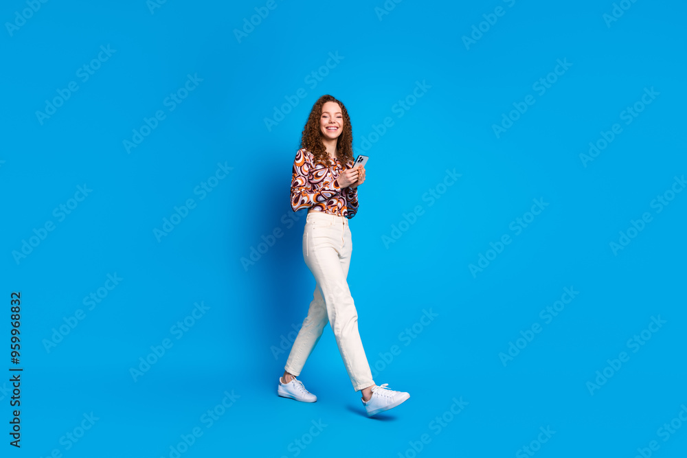 Full length photo of lovely young lady hold device walking dressed stylish colorful garment isolated on blue color background Full length photo of lovely young lady hold device walking dressed stylish colorful garment isolated on blue color background