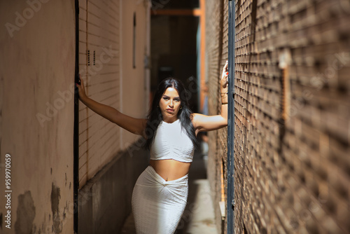 Young, beautiful, brunette, Latina woman in a white suit, posing on a very narrow street leaning against the wall, receiving the sun's rays. Concept beauty, fashion, trend, city. photo