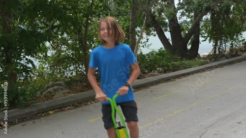 A young boy on a green pogo stick plays joyfully by the seaside, near rocks and trees. The tranquil ocean and a distant pier create a serene and adventurous atmosphere photo