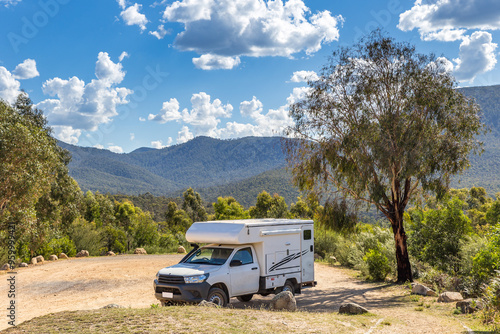 A camper van is parked beneath a large tree at a campsite surrounded by lush greenery and mountains, with fluffy clouds scattered in the blue sky.