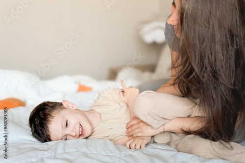 A young mother in casual clothes plays tickle with her happy little son. Mom and child have fun together in a cozy bedroom at home. Happy children with mother.