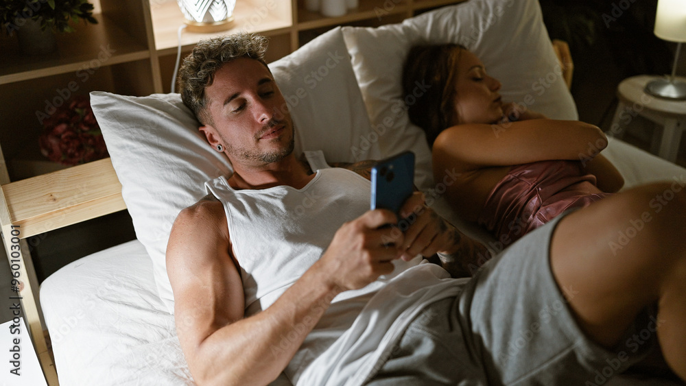 Obraz premium A man and woman share a quiet moment in their bedroom, with the man browsing his smartphone and the woman sleeping beside him.