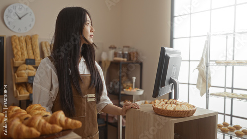 Sticker Young chinese woman working in a bakery, standing near the counter indoors with various bread displays around her and a sunny interior with a clock on the wall in china