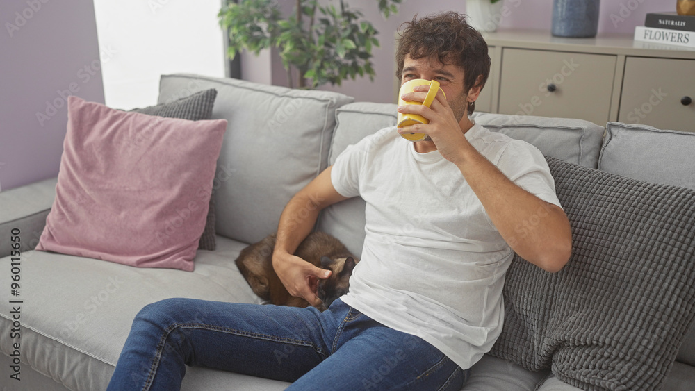 A relaxed young hispanic man enjoying a beverage while petting a siamese cat on a couch in a cozy living room. A relaxed young hispanic man enjoying a beverage while petting a siamese cat on a couch in a cozy living room.
