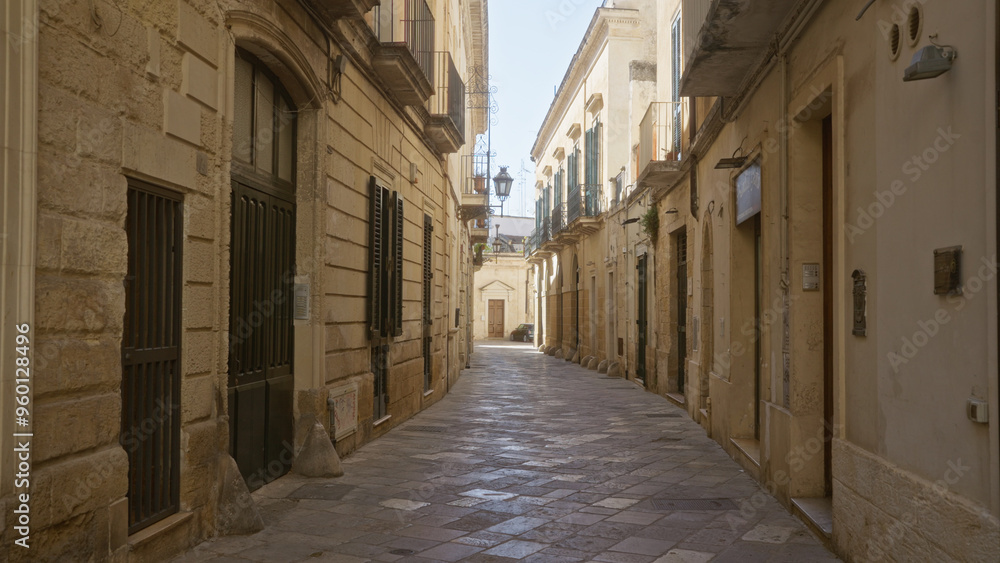 Fototapeta premium A quiet, narrow street in lecce, puglia, italy, showcasing historic architecture and mediterranean charm with closed shutters and cobblestone paving on a sunny day.