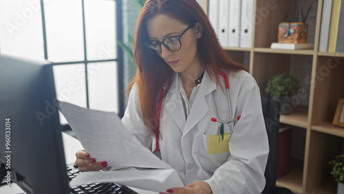 Canvas-taulu A focused redhead woman in a white coat analyzes a document in a clinic office, showcasing healthcare and professionalism