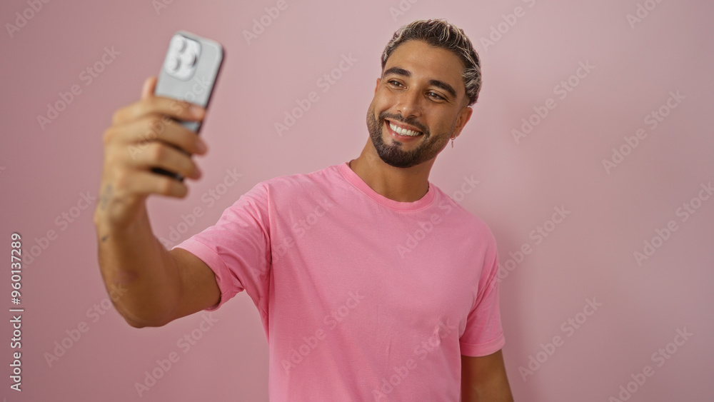 Handsome young muslim man with a beard, wearing a pink t-shirt, takes a selfie against a pink isolated background, smiling joyfully. Handsome young muslim man with a beard, wearing a pink t-shirt, takes a selfie against a pink isolated background, smiling joyfully.