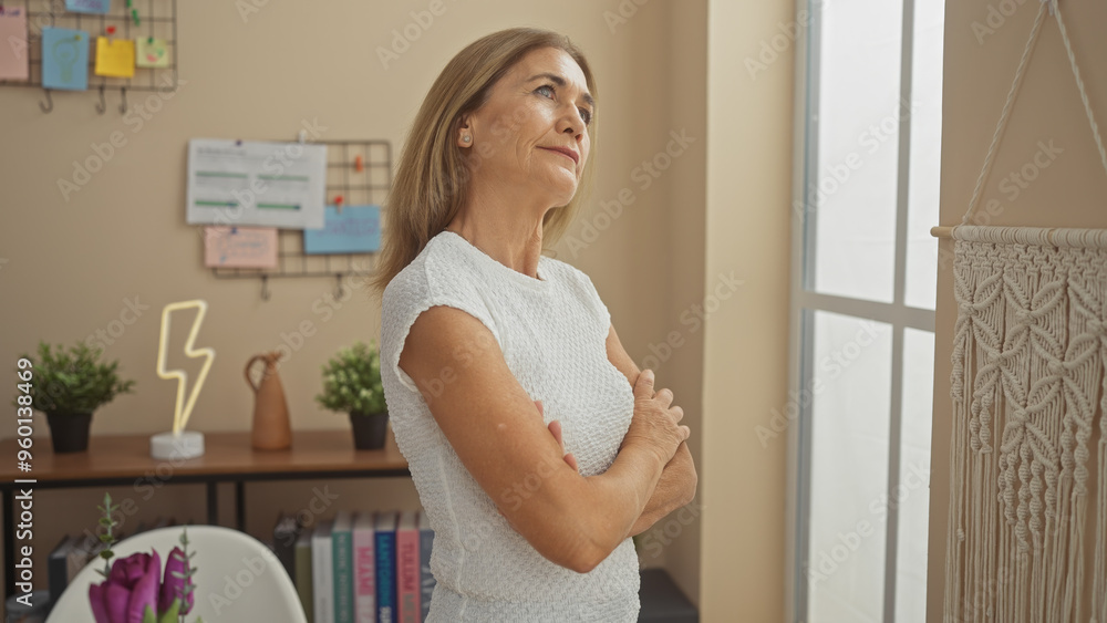 A contemplative middle-aged woman stands with folded arms in a cozy living room, surrounded by modern decoration and sunlight. A contemplative middle-aged woman stands with folded arms in a cozy living room, surrounded by modern decoration and sunlight.