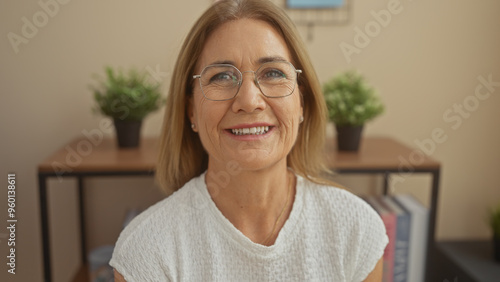 A smiling middle-aged woman with glasses wearing a white sweater in a home interior setting. Free Download A smiling middle-aged woman with glasses wearing a white sweater in a home interior setting. - GFXLoad