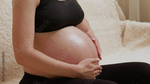 Close-up image of a pregnant woman in her last trimester sitting on the couch massaging her belly with moisturizing oil to prevent stretch marks during pregnancy. Self care during pregnancy photo