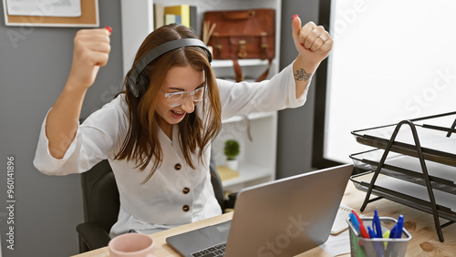 Obraz na plátně Excited woman with headphones celebrating success at modern office workplace