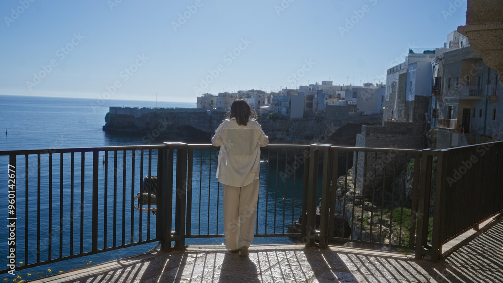 A young hispanic woman enjoys a stunning view from a viewpoint overlooking the tranquil water and historic buildings in polignano a mare, puglia, italy on a sunny day. A young hispanic woman enjoys a stunning view from a viewpoint overlooking the tranquil water and historic buildings in polignano a mare, puglia, italy on a sunny day.