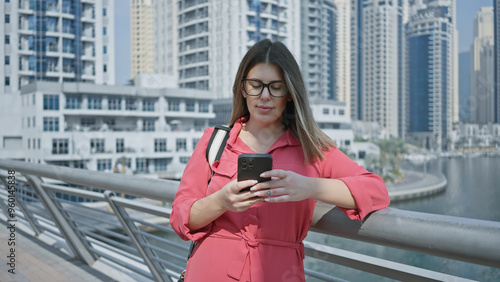 Canvas Print A stylish woman in glasses uses a smartphone against the backdrop of dubai marina's luxury skyline