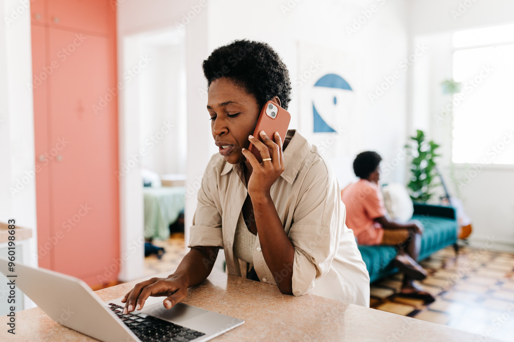 Working mom speaking on the phone while using a laptop in a home kitchen Working mom speaking on the phone while using a laptop in a home kitchen